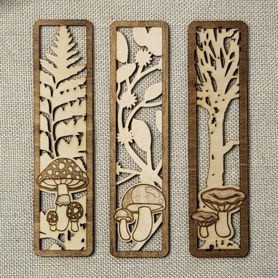 Three wooden bookmarks with laser-cut nature designs on a textured fabric background