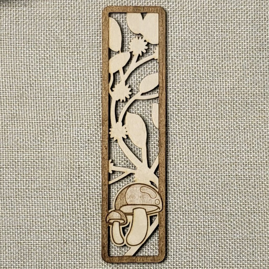 Wooden bookmark with laser-cut design of mushrooms and foliage on a textured fabric background.