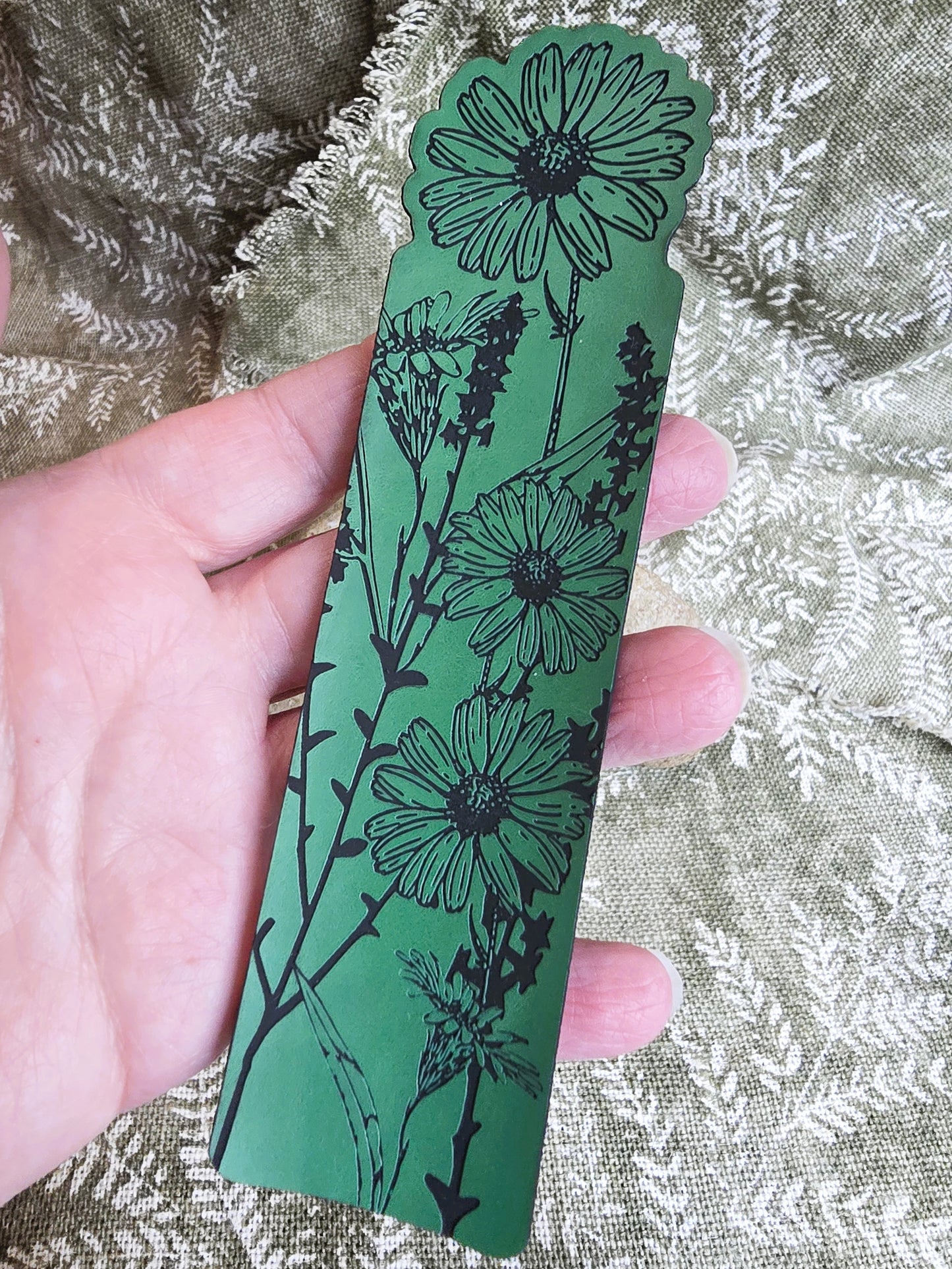 Hand holding a green bookmark with black floral design on a textured fabric background