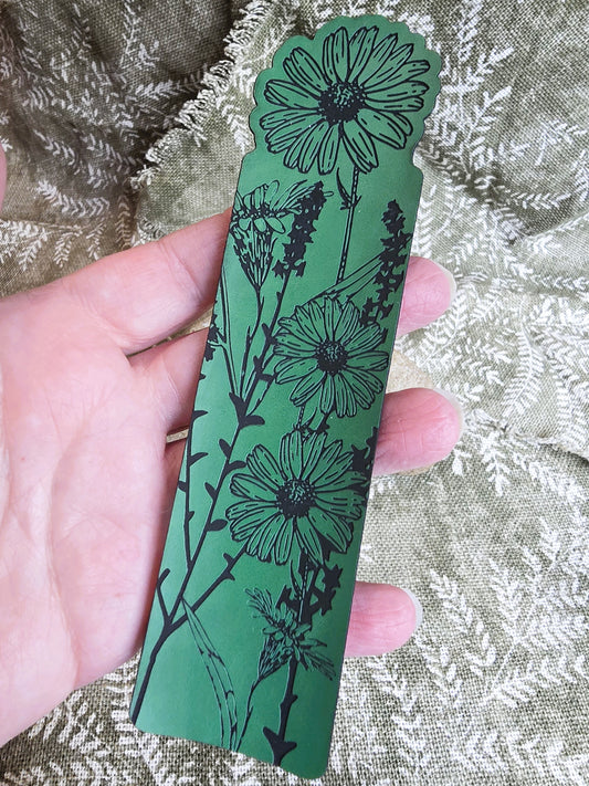 Hand holding a green bookmark with black floral design on a textured fabric background